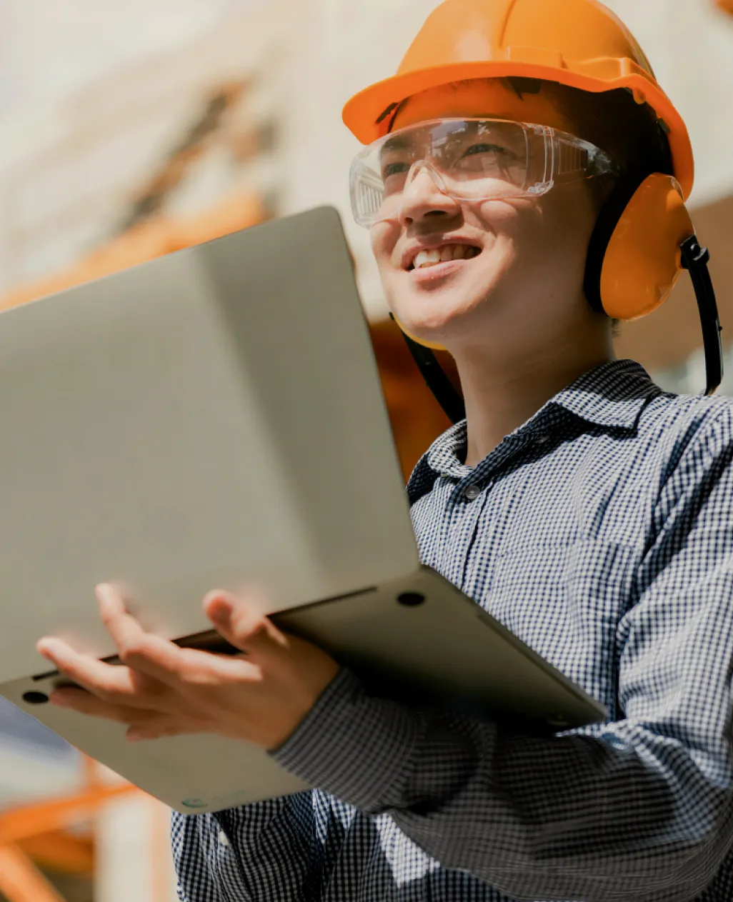 A man wearing a safety helmet and glasses holding a laptop in a construction zone