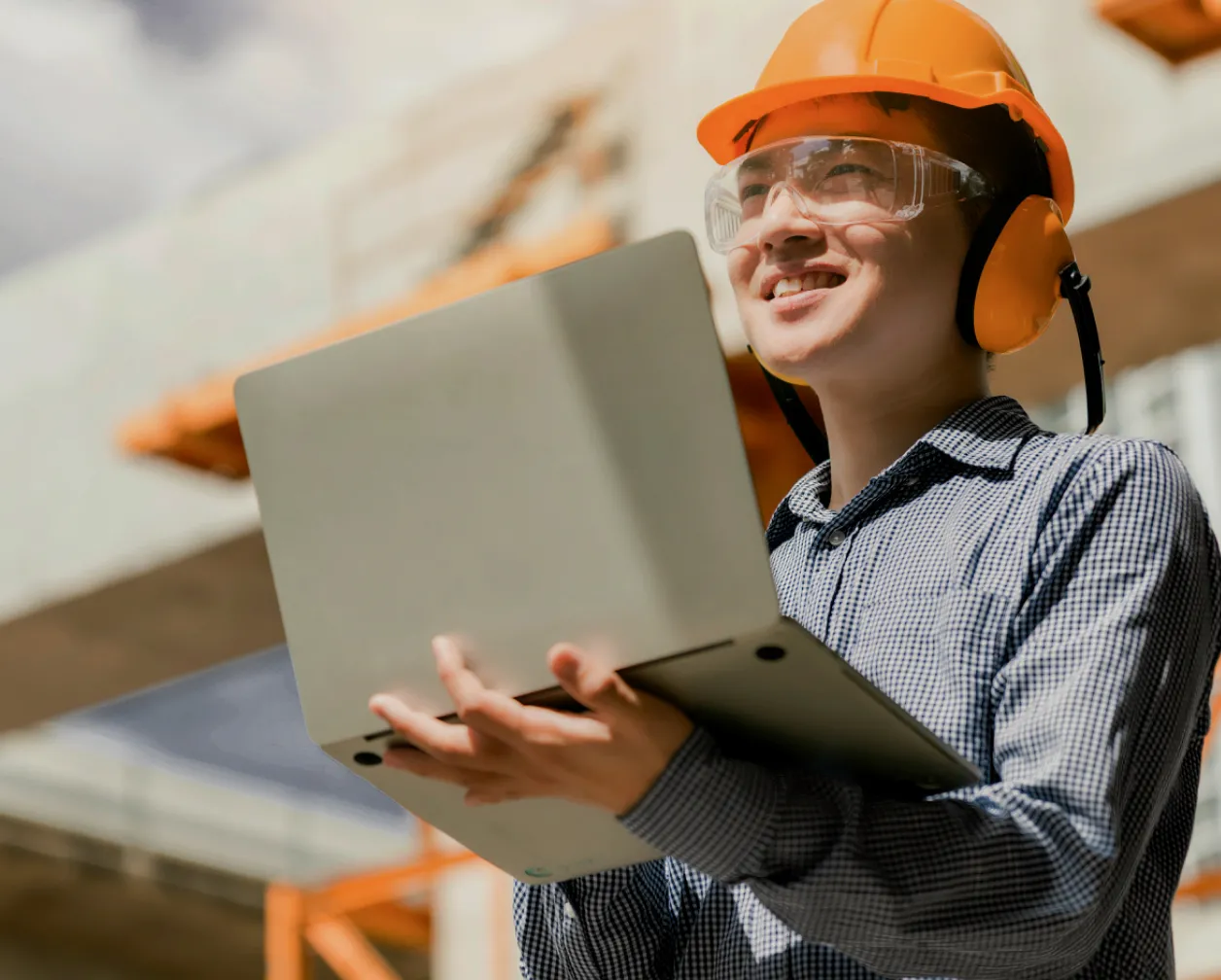 A man wearing a safety helmet and glasses holding a laptop in a construction zone