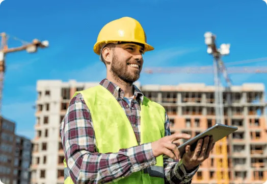 A man wearing a hard hat and safety vest is holding a tablet computer on a construction site.