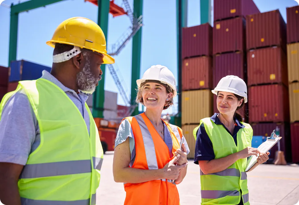 Two women in hard hats and safety vests discuss with an engineer besides cargo containers at a construction site.